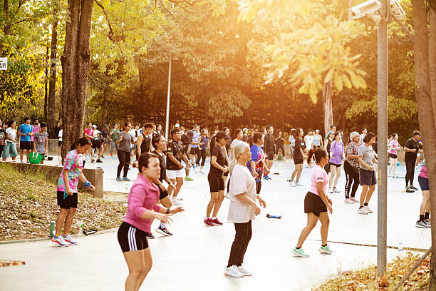 Large group of thai people is doing aerobic in Wachirabenchatat Park  in Bangkok at late afternoon with deep standing sun over treetops. People are exercising on small rounded square surrounded by trees near anrtrance of park from Kampheng Phet Road. There are many adult and mature adult women and men following an instructor and music at right side on stage out of frame