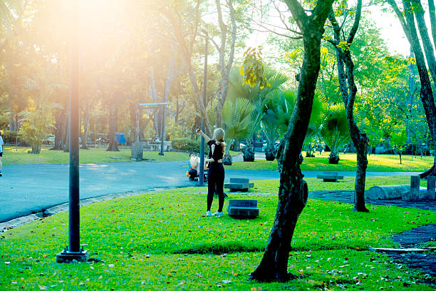 Thai woman is taking selfie in early sunset light of deep sun over treetops in Chatuchak Park in Bangkok. Woman is wearing black leggings and spports shoes. She has blond colored hair and is standing among trees. Scene is in northern area of the park. Behind woman a person is squatting  at a baby stroller and is watching blond haired woman