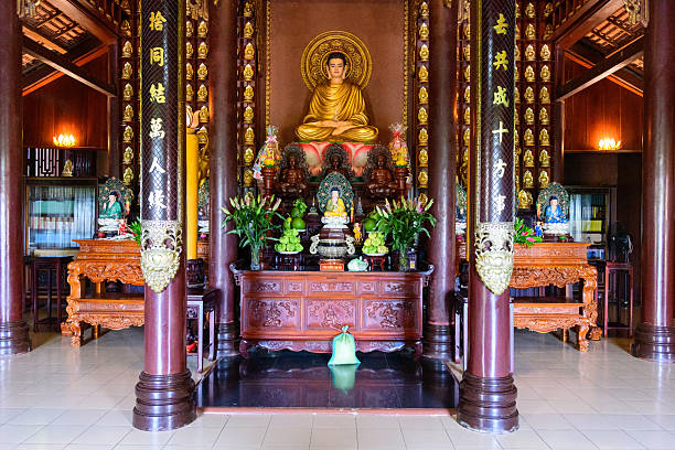Elaborate Buddhist altar featuring a golden statue, intricate wood carvings, and decorative columns inside a traditional temple hall. The space is filled with vibrant offerings, lush green plants, and warm ambient lighting.