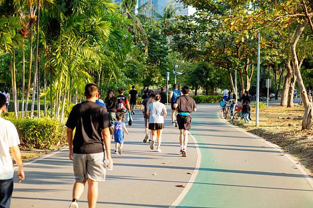 Walking and jogging thai people and families in Wachirabenchatat Park  in Bangkok. Park is also called Rot Fai Park.. People are on one of major pathways. Some are wearing sports clothes and are running. Rear view and capture of people. Among adults is a little boy in sports clothes. At right side is cycling lane. There some young muslim thai women are standing with bicycles.