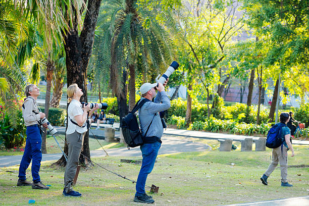 White mature adults and photographers with cameras and large tle zoom lenses captured in Wachirabenchatat Park  in Bangkok. Park is also called Rot Fai Park.. There are two men and  one woman. People are watching and focusing on tropical birds up in treetops.One man is wearing a backback. In right are people are walking