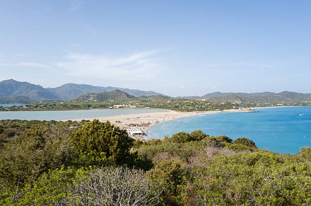 Expansive sandy shoreline curves between a turquoise sea and a calm lagoon at Porto Giunco, backed by lush hills and distant mountains under a clear sky.