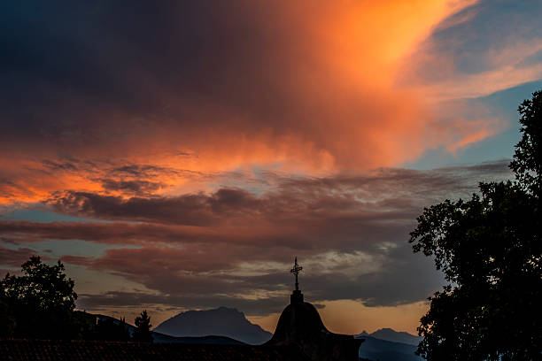 Dramatic sunset sky with glowing clouds above a church cross and mountain silhouettes. Powerful Mediterranean evening scene in Corte, Corsica, France, expressing emotion, contrast and spiritual atmosphere.
