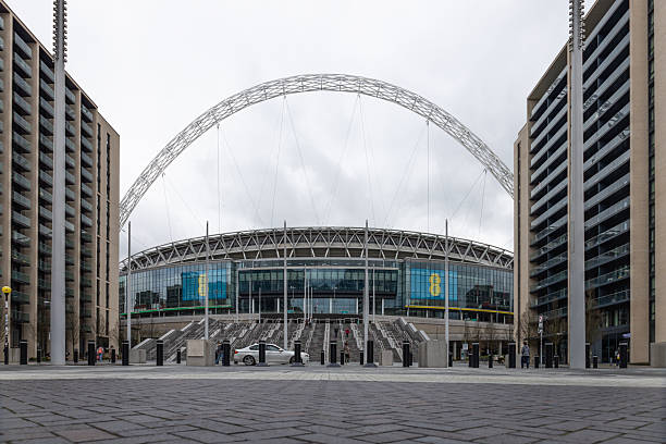 A wide view of the iconic Wembley Stadium arch and main entrance framed by modern apartment buildings under a cloudy sky. London, UK, 17 March 2024
