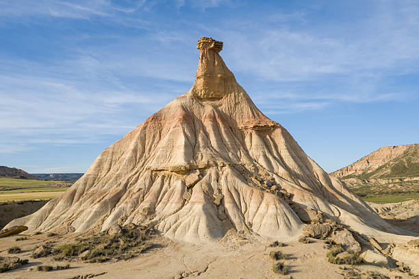 A dramatic hoodoo with sculpted beige slopes rises from the arid landscape in Bardenas Reales, Spain. Sunlight casts sharp shadows, revealing rugged textures and layered earth beneath a clear blue sky.