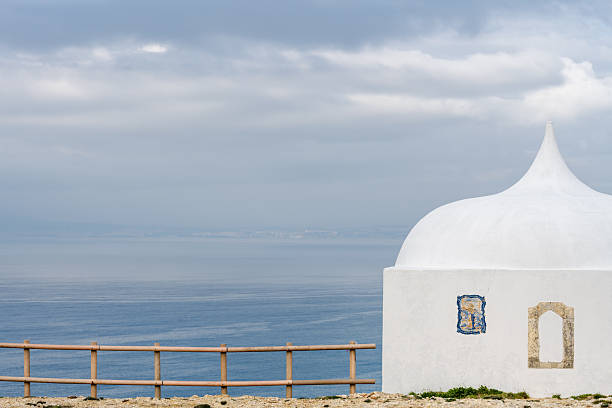 Sesimbra Portugal December 25 2025. A close view shows the white dome of a coastal chapel with a wooden fence and the Atlantic Ocean behind at Cabo Espichel