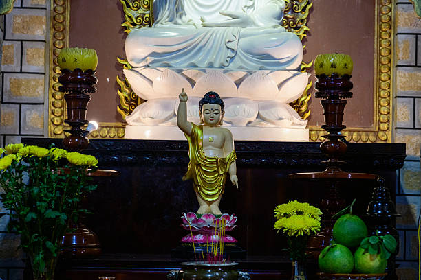 Golden baby Buddha statue stands on a pink lotus pedestal at the center of a richly decorated altar, surrounded by yellow flowers, fruit offerings, and dark wood candle holders. Warm lighting highlights the intricate details and serene spiritual ambiance.