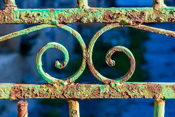 A close-up of a green, rusted wrought-iron railing featuring curled spirals and peeling patina, suggesting vintage charm and weathered outdoor architecture.