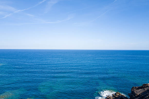 Expansive view of the tranquil blue Mediterranean Sea stretching to the horizon, with gentle waves and a rocky shoreline in the foreground under a clear, sunlit sky near Argentiera, Sardinia.