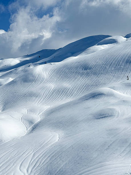 Pristine snowy alpine slopes shaped by wind and early spring rainfall, revealing flowing lines and layered textures. Soft sunlight, shadows and passing clouds in remote winter mountain landscape.