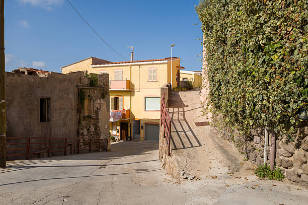 Quiet sunlit street in the historic center of Castelsardo, featuring pastel buildings, textured stone walls, and lush green ivy under a clear blue sky.