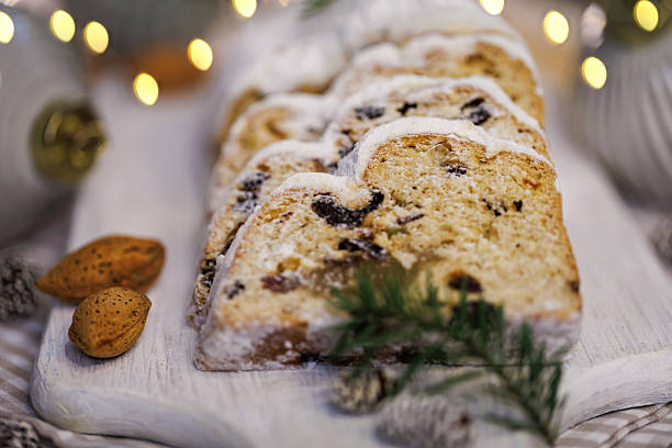A beautifully sliced Christmas stollen dusted with powdered sugar, accompanied by almonds, pinecones, and festive decorations on a wooden board, with blurred warm holiday lights.