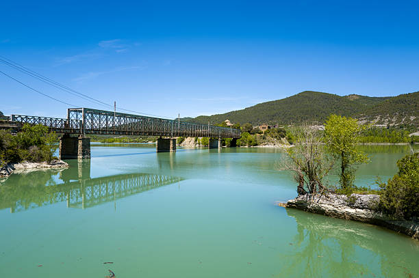A steel truss bridge spans calm turquoise water with reflections, framed by rocky shores, green trees, and distant forested hills under a vivid blue sky at Las Penas.