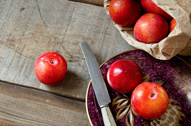 Red plums spill from a crumpled paper bag onto rustic wood near a knife and ornate plate, with generous copy space around the fruit.