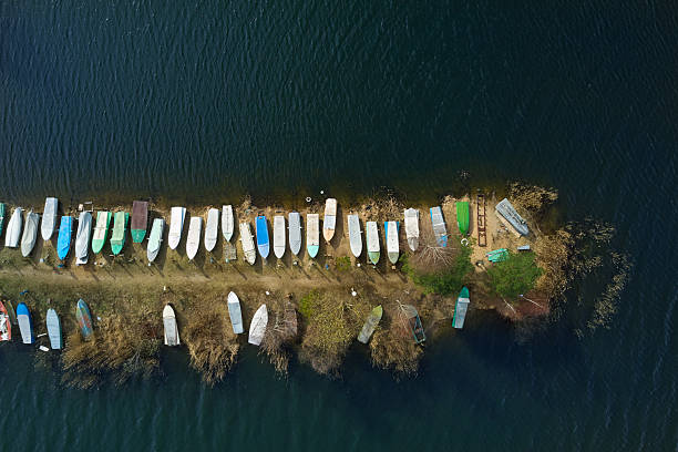 Aerial top-down view of small boats pulled out onto the river shore and arranged along the shoreline. Calm water surface, natural riverbank with vegetation, and a row of colorful boats resting on land. Concept of fishing, water transport, outdoor recreation, and peaceful nature.