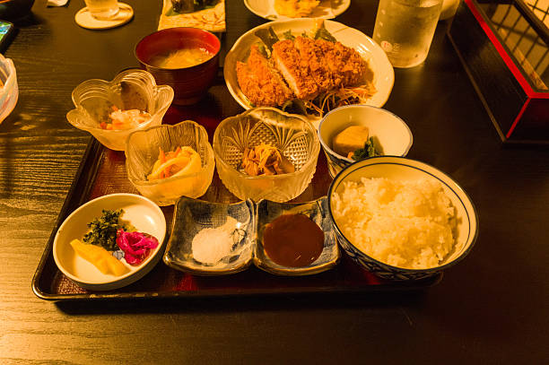 Traditional Japanese meal set featuring steamed white rice, fried breaded cutlet, pickled vegetables, and assorted small side dishes on a lacquered tray under warm ambient lighting in Hongu, Japan.