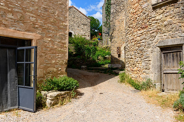 Quiet gravel lane in La Couvertoirade bordered by textured stone buildings, open blue-framed door, weathered wooden doorway, and lush greenery under a bright summer sky.
