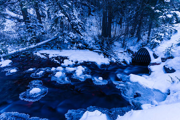 Scenic creek with fresh snow and ice with flowing water going through corrugated steel culvert under a road.