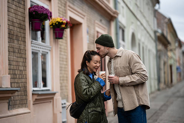 Young couple expressing affection on an urban street, man kissing woman's forehead while holding coffee cups