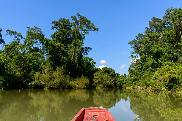 A red wooden boat drifts on a calm river surrounded by dense tropical forest and lush greenery near Konglor Cave in central Laos. Clear blue sky and vivid reflections create a tranquil scene in this remote limestone region.