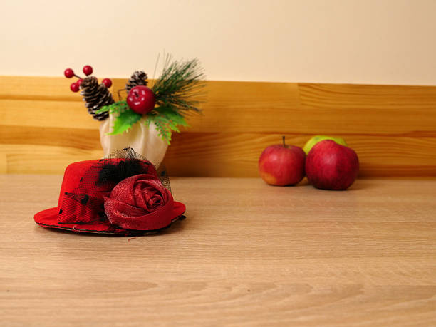 Red decorative hat with flower on table, apples and seasonal decor in background.