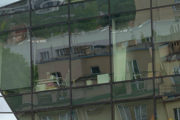 glass facade reflecting warped urban scene tilted window panels distort neighboring rooftops, trees and interior chairs into layered abstract shapes, creating modern