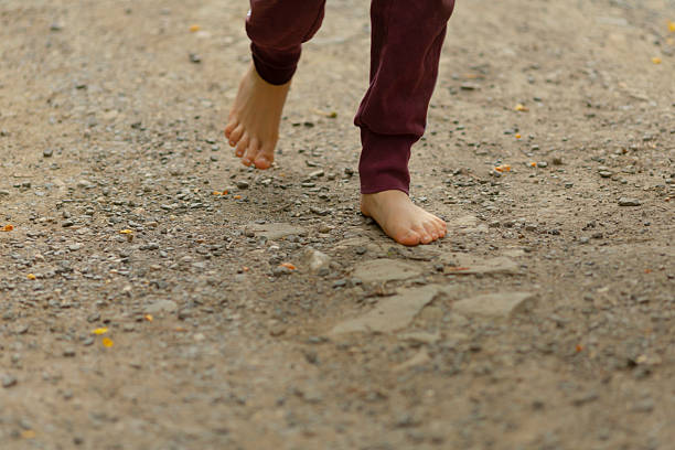 barefoot child walking on gravel path midstep closeup showing toes and soles, maroon sweatpants cuffed at ankle, scattered pebbles and dirt underfoot, candid outdoor play