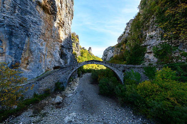 Arched stone bridge spans a dry rocky riverbed between towering limestone cliffs and dense autumn foliage in the Vikos Gorge region of Epirus, Greece. Soft daylight reveals textured masonry and vibrant natural surroundings in this dramatic mountain landscape.