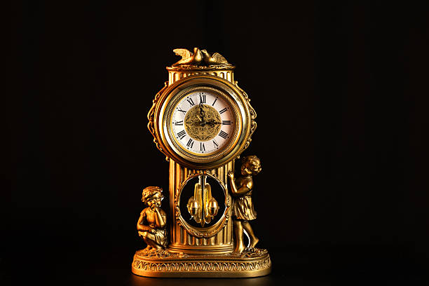 A close-up studio shot of an ornate, antique golden mantel clock with cherub figures on either side, set against a dark background.
