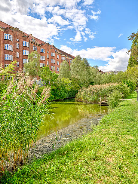 View of a serene urban area featuring brick buildings reflecting historical charm, dense greenery surrounding a pond, a pier, and sunny weather. The location reflects a peaceful corner in Denmark.