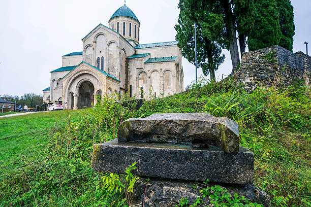 A wide-angle view of the historic Bagrati Cathedral (The Cathedral of the Dormition) in Kutaisi, Georgia, on a cloudy day. The image features the medieval stone architecture with its controversial modern restoration elements, a historical information board showing the reconstruction process, and the surrounding stone ruins and greenery. An iconic landmark representing Georgian Golden Age architecture.