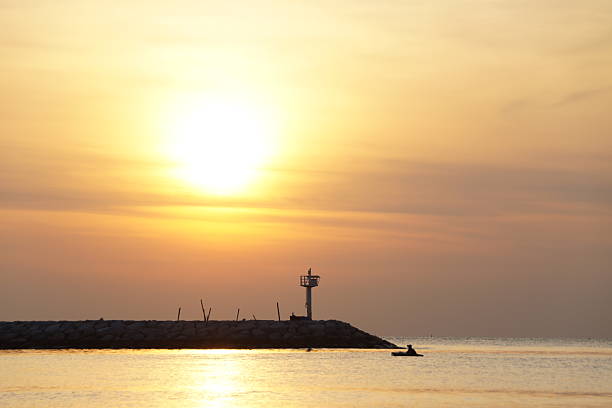 Golden sunrise at sea, stone pier, lighthouse and tourist on a board.