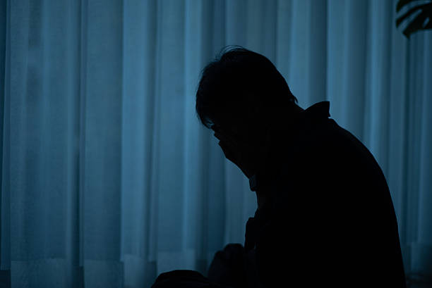 Silhouette of a lonely man sitting by a window at night feeling depressed.