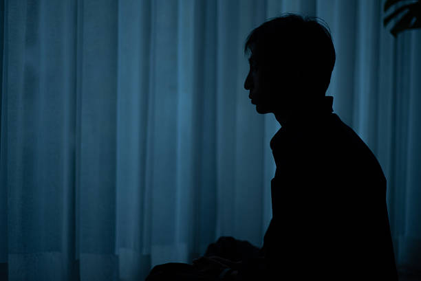 Silhouette of a lonely man sitting by a window at night feeling depressed.