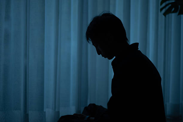 Silhouette of a lonely man sitting by a window at night feeling depressed.