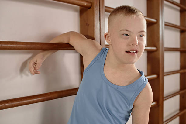 Caucasian boy child with Down syndrome exercising on gym wall bars, wearing sleeveless top, looking away from camera, showing active participation in physical activity