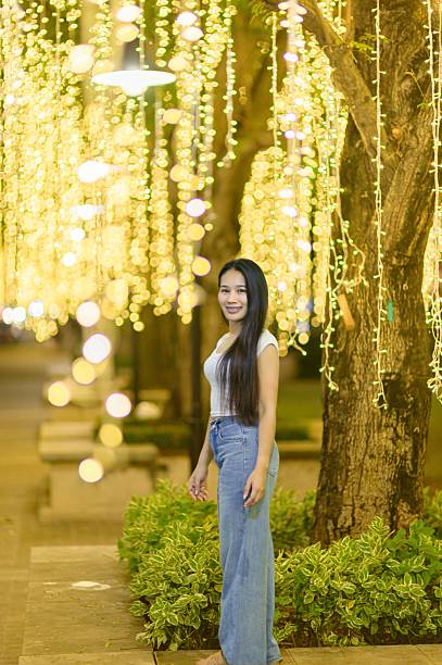 A joyful woman stands gracefully amidst a backdrop of glowing decorative lights, creating a warm and inviting ambiance during the evening in an outdoor setting.