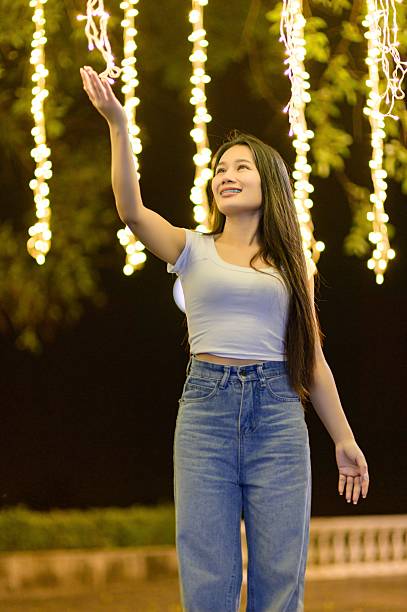 A young woman joyfully reaches out toward sparkling light bulbs hanging in a magical outdoor setting at night, creating a vibrant and cheerful atmosphere.