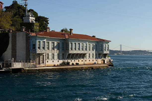 Istanbul, Turkey-January 23, 2026:  A scenic view of a traditional Ottoman-style wooden mansion, known as a Yali, located directly on the shoreline of the Bosphorus. The 15 July Martyrs Bridge (formerly Bosphorus Bridge) is visible in the distance under a clear blue sky, connecting the European and Asian sides of the city.