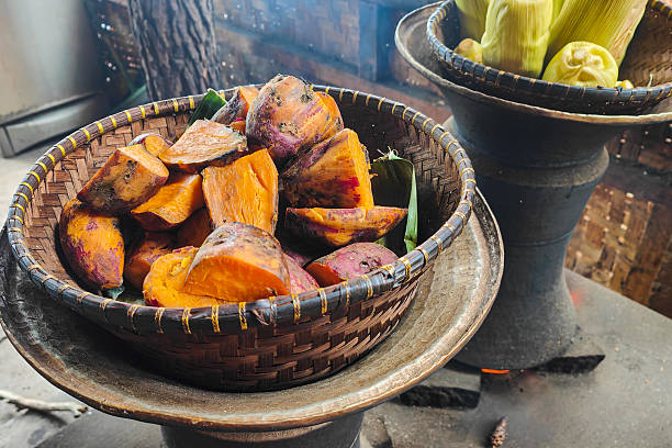 Close up shot of freshly steamed sweet potatoes and corn arranged in a woven bamboo basket, placed over a traditional charcoal stove in a rustic kitchen setting.ConceptSweet potatoes, traditional cooking, steamed food, local cuisine, rustic kitchen, bamboo basket, Asian food, healthy carbohydrate, home cooking, traditional stove
