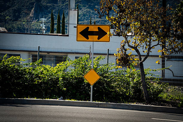 A yellow road sign with black arrows pointing left and right, indicating two-way traffic, stands beside a lush green bush and a tree