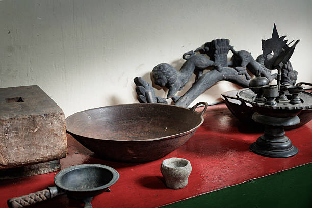 Traditional Indonesian kitchenware and ritual objects including metal wok, stone mortar, wooden tools, and carved decorative elements displayed on a rustic wooden table, representing cultural heritage and traditional lifestyle