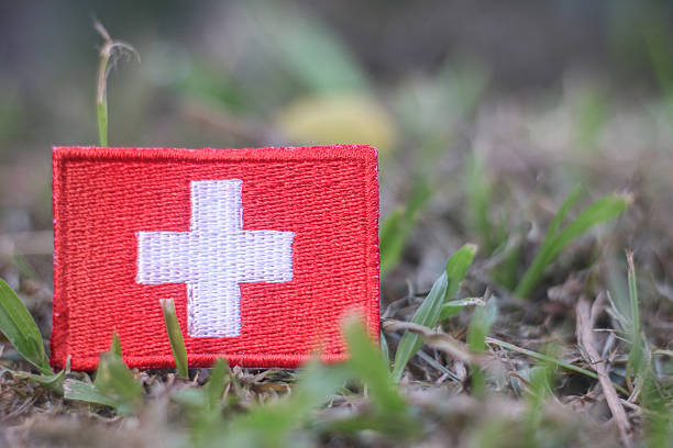 An embroidered Swiss flag patch is positioned in natural green grass with a soft blurred outdoor background. The image symbolizes Switzerland national identity combined with environmental awareness, sustainability, and ecological responsibility. It reflects themes of conservation, earth day, climate awareness, rural landscapes, and the balance between cultural identity and respect for nature. Suitable for editorial, educational, and environmental communication.