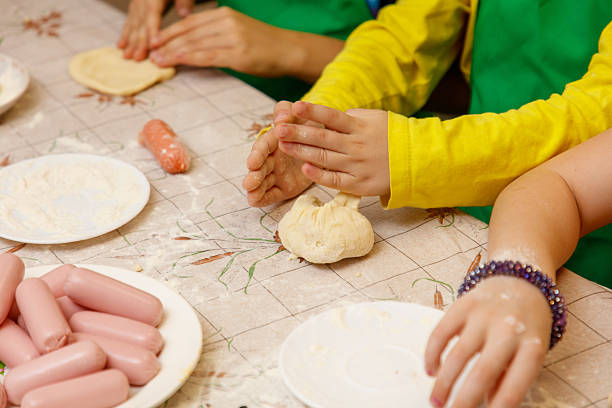 A group of children are making dough on a table. There are several plates of dough on the table, and a few hot dogs are on the table as well