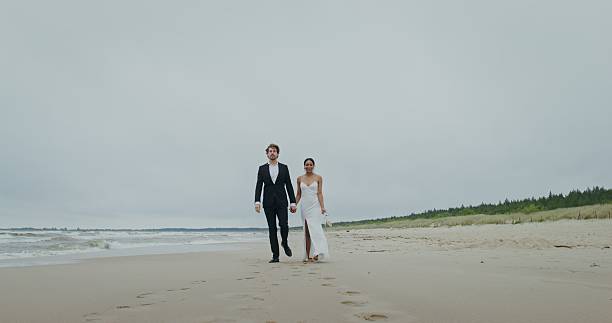 The bride and groom hold hands walking on the beach, the bride holds shoes in her hands