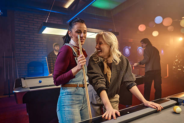 Two women share a joyful moment by a pool table in a vibrant, neon arcade bar, while others play in the background, creating a lively, social atmosphere.