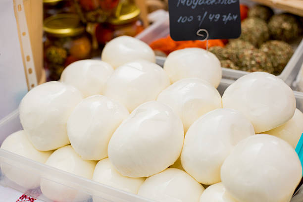 Pile of smooth white mozzarella cheese balls in plastic container with handwritten price tag and pickled vegetables in background
