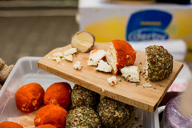 Artisan cheese balls coated in red paprika and green herbs displayed on cutting board with plastic containers showing specialty varieties
