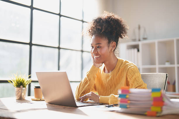istock Young woman with curly hair smiling at her laptop while working or studying at a bright modern desk in a sunlit home office, focused and productive 2256026844