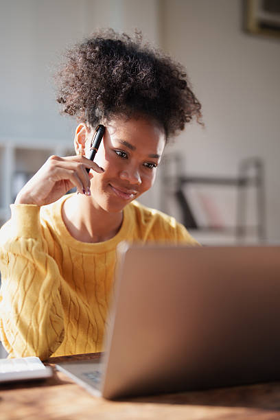 Young adult woman actively thinking and collaborating online, holding a pen and looking at a laptop screen in a bright home office environment, representing remote work and education